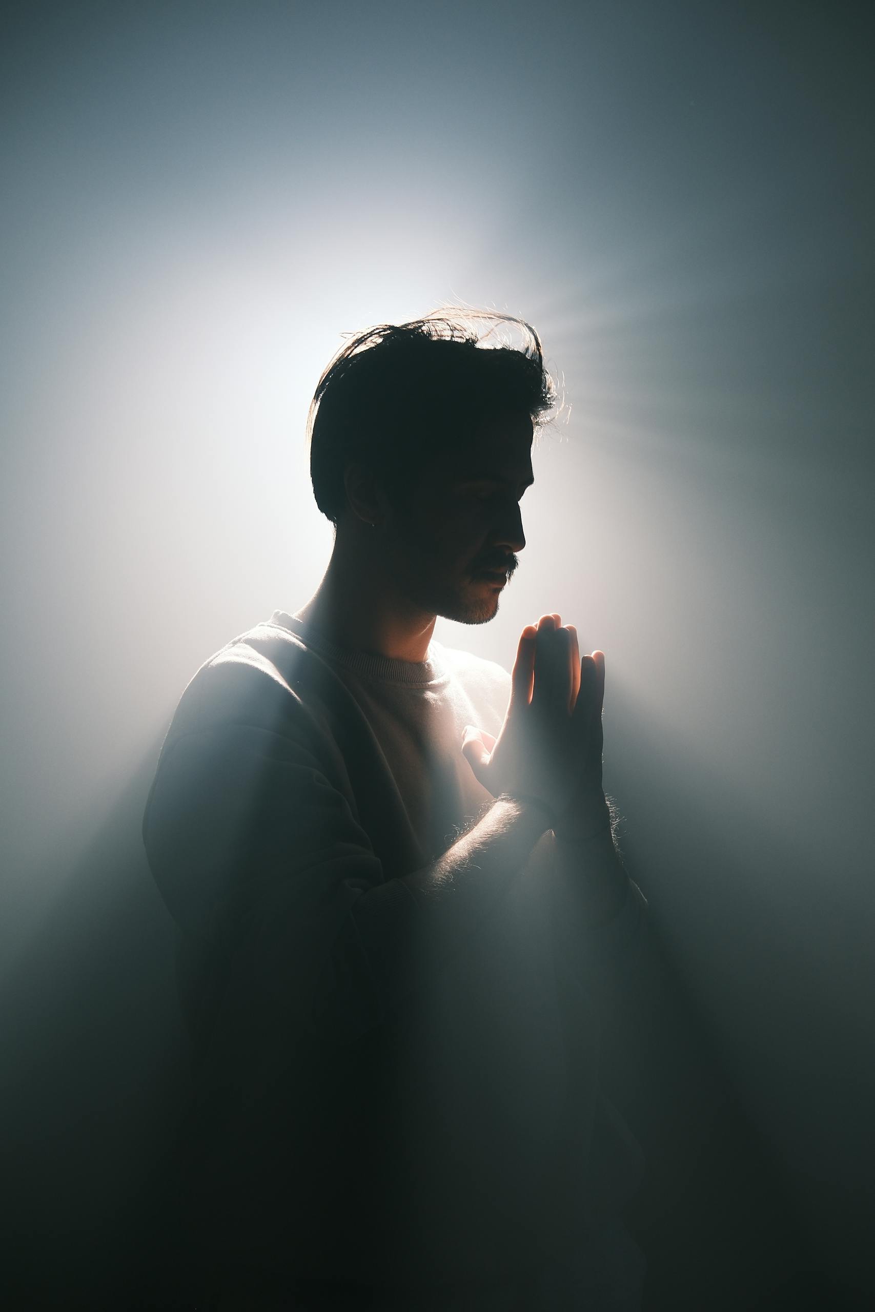 A silhouette of a man in prayer with dramatic backlighting creating a halo effect and light rays.