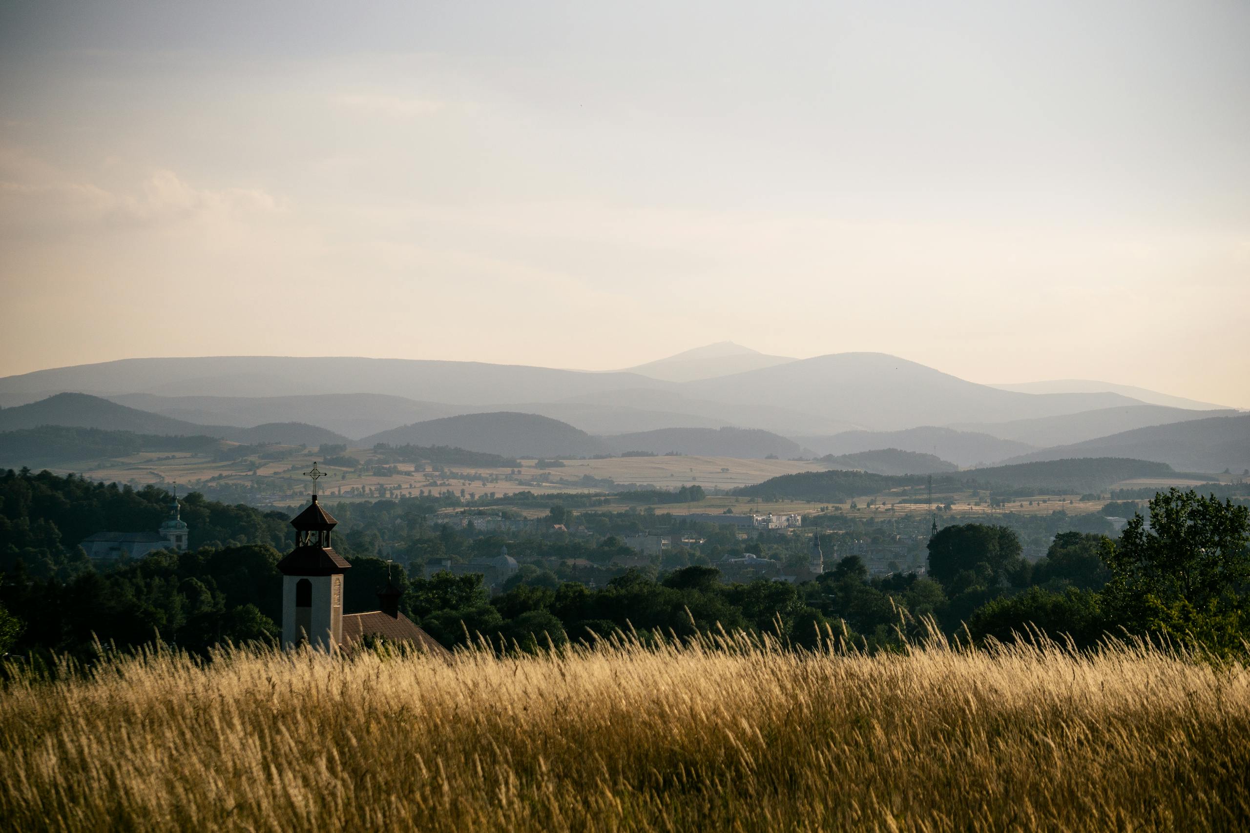 Tranquil landscape of Kamienna Góra, Poland, featuring lush fields, hills, and a distant church under a sunset sky.