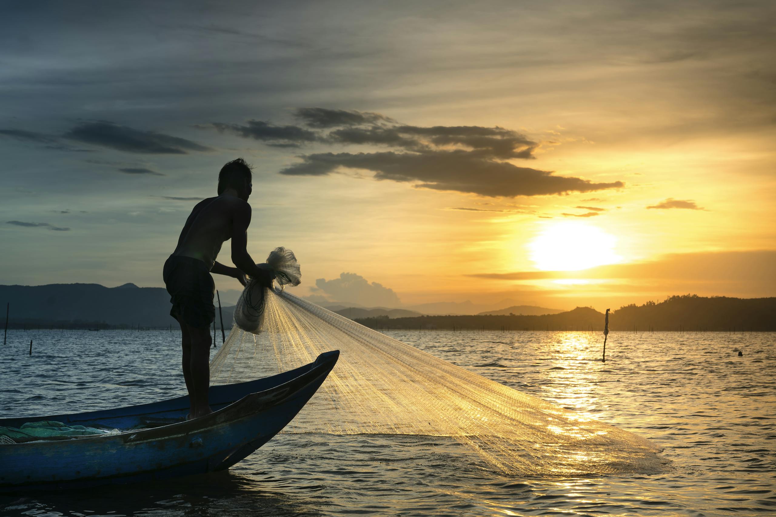 Silhouette of a fisherman casting a net from a boat at sunrise on a tranquil lake.